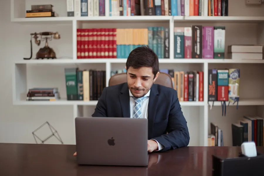 Ao notebook, à mesa de trabalho, diante da mesma estante de livros jurídicos.