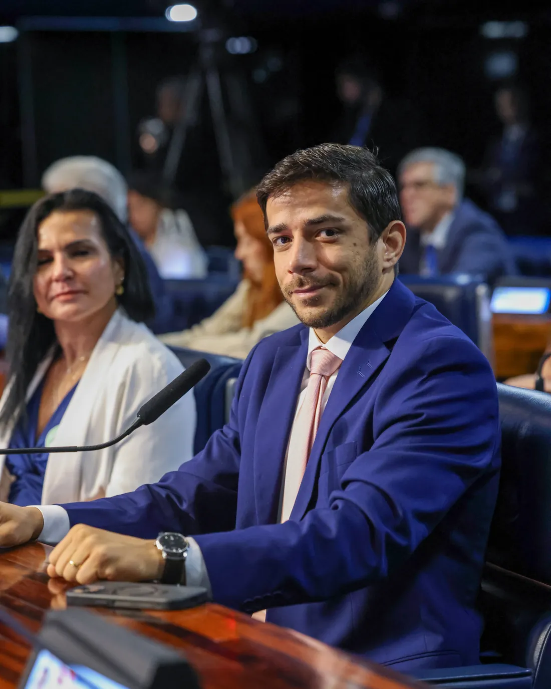 O Dr. Victor Hugo Passos Ferreira, de terno azul, sentado à bancada de madeira e olhando para a câmera, com a Dra. Ivone Duque ao lado.
